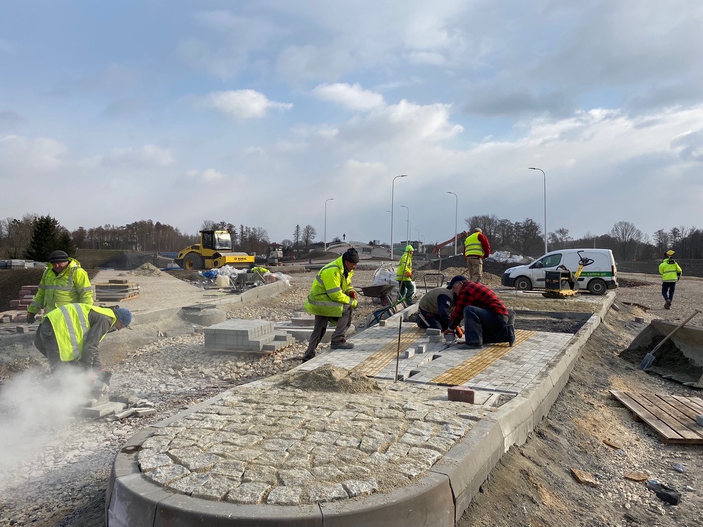 Ekipa brukarska w trakcie układania kostki na placu budowy. Widoczny walec drogowy i samochód dostawczy. Praca wre w pochmurny dzień.