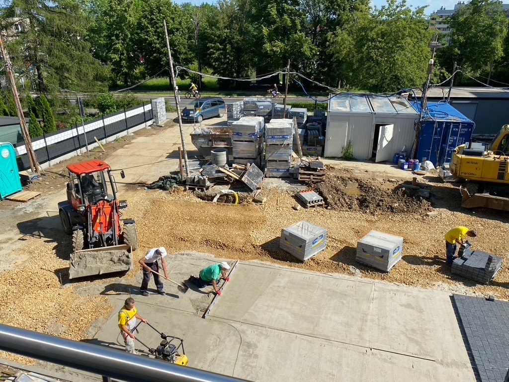 Intensywne prace brukarskie: robotnicy wyrównują podłoże, ubijają ziemię i układają kostkę. Na placu budowy stoi traktor i palety z kostką. Widok z góry.