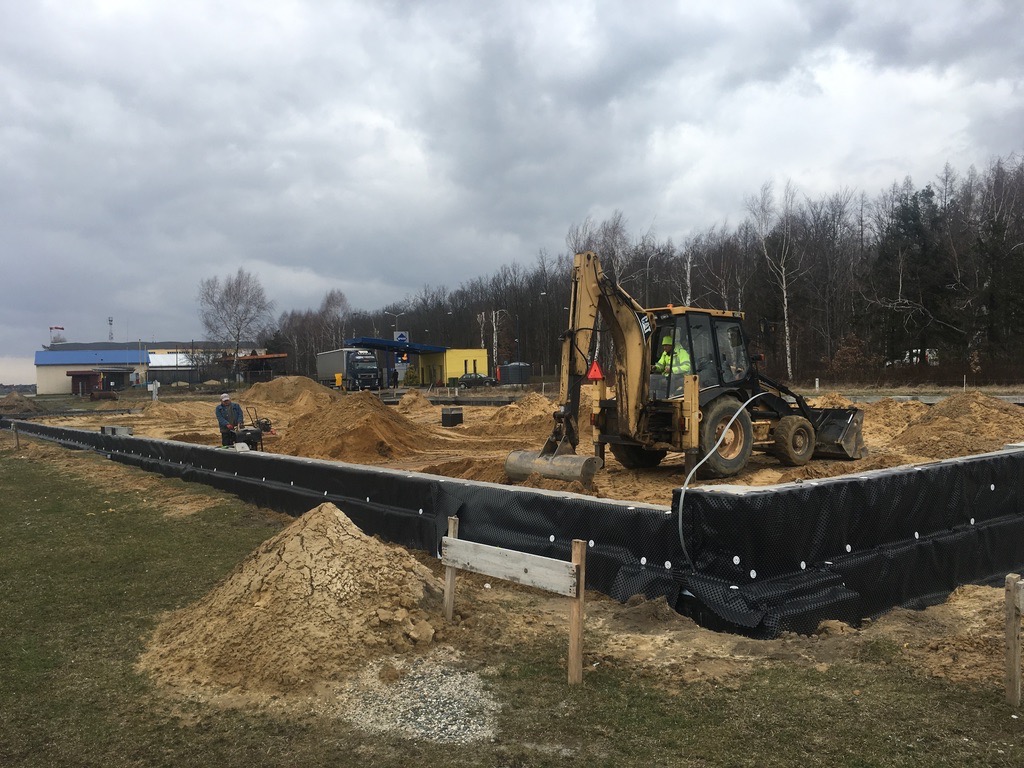 Żółta koparko-ładowarka w wykopie z zabezpieczonymi ścianami. W tle operator zagęszczarki. Prace ziemne w toku, pochmurne niebo.