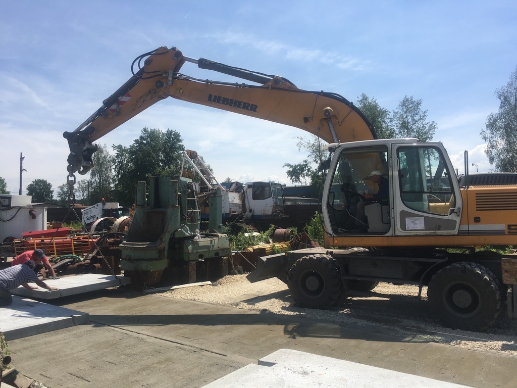 Żółta koparka Liebherr podnosi betonową płytę. Dwóch robotników pomaga ustawić płytę. W tle widać inne maszyny i drzewa pod błękitnym niebem.