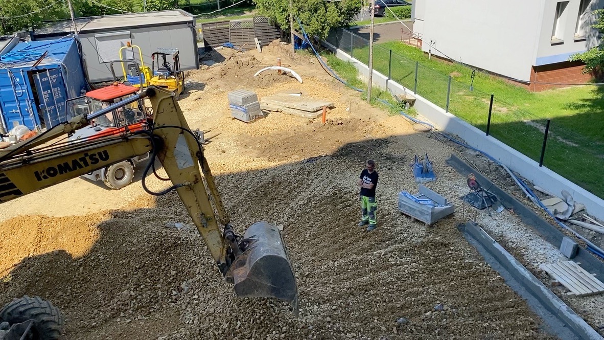 Żwirowe podłoże wyrównane koparką Komatsu na budowie. Widoczne palety z kostką, fundament, ogrodzenie panelowe i pracownik w tle.