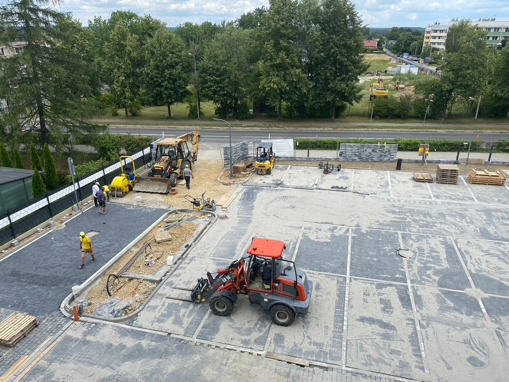 Układanie kostki brukowej na parkingu. Widoczny sprzęt budowlany i pracownicy. Część parkingu już wyłożona kostką, reszta przygotowana pod ułożenie. Praca w toku.