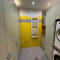 BEE PERFECT - Funkcjonalna pralnia z szafami na wymiar w odcieniach szarości i żółci. Widoczna pralka Bosch, grzejnik drabinkowy i płytki ceramiczne na ścianie.