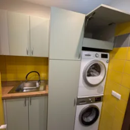 Funkcjonalna zabudowa: pralka i suszarka w szafie na wymiar z szafkami kuchennymi w pistacjowym odcieniu, żółte płytki na ścianie i szara podłoga.