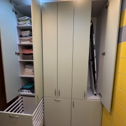 BEE PERFECT - Jasna szafa na wymiar z otwartymi drzwiami, wewnątrz półki z ręcznikami i deską do prasowania. Dolna część z wysuwanym koszem. Żółto-szara ściana w tle.