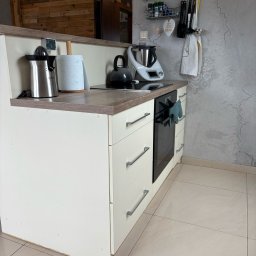 BEE PERFECT - Fragment kuchni z białymi szafkami, drewnianym blatem i sprzętami AGD, w tle ściana z szarą strukturą i drewniane panele. Widok z perspektywy.