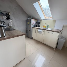 BEE PERFECT - Jasna kuchnia na poddaszu z oknem dachowym. Kremowe szafki, drewniany blat, srebrna lodówka z magnesami i szara ściana z dekoracyjną fakturą.