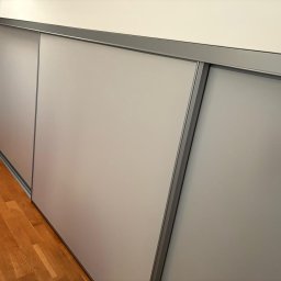 BEE PERFECT - Nowoczesna szafa wnękowa z przesuwnymi, matowymi drzwiami w kolorze szarym. Widoczny fragment drewnianej podłogi i metalowe prowadnice. Minimalistyczny design.