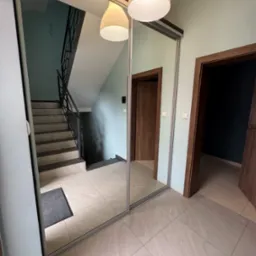 Szafa wnękowa z lustrzanymi drzwiami przesuwnymi w przedpokoju, odbijająca schody i lampę sufitową. Widoczne dwie pary drzwi po bokach szafy.