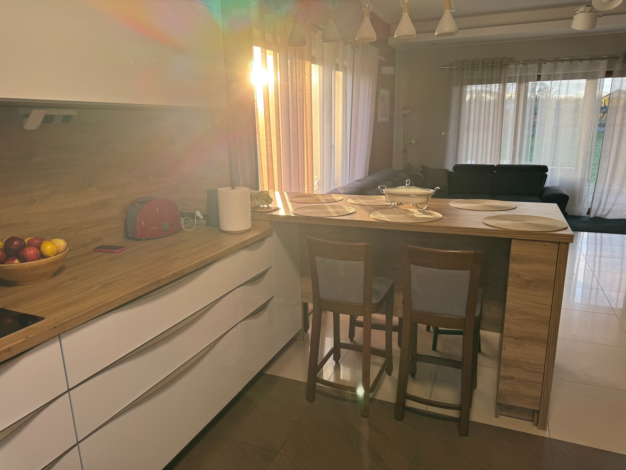 Jasna kuchnia z drewnianym blatem i białymi frontami, wysoki barek z krzesłami. W tle okno z firankami przepuszczające promienie słoneczne.