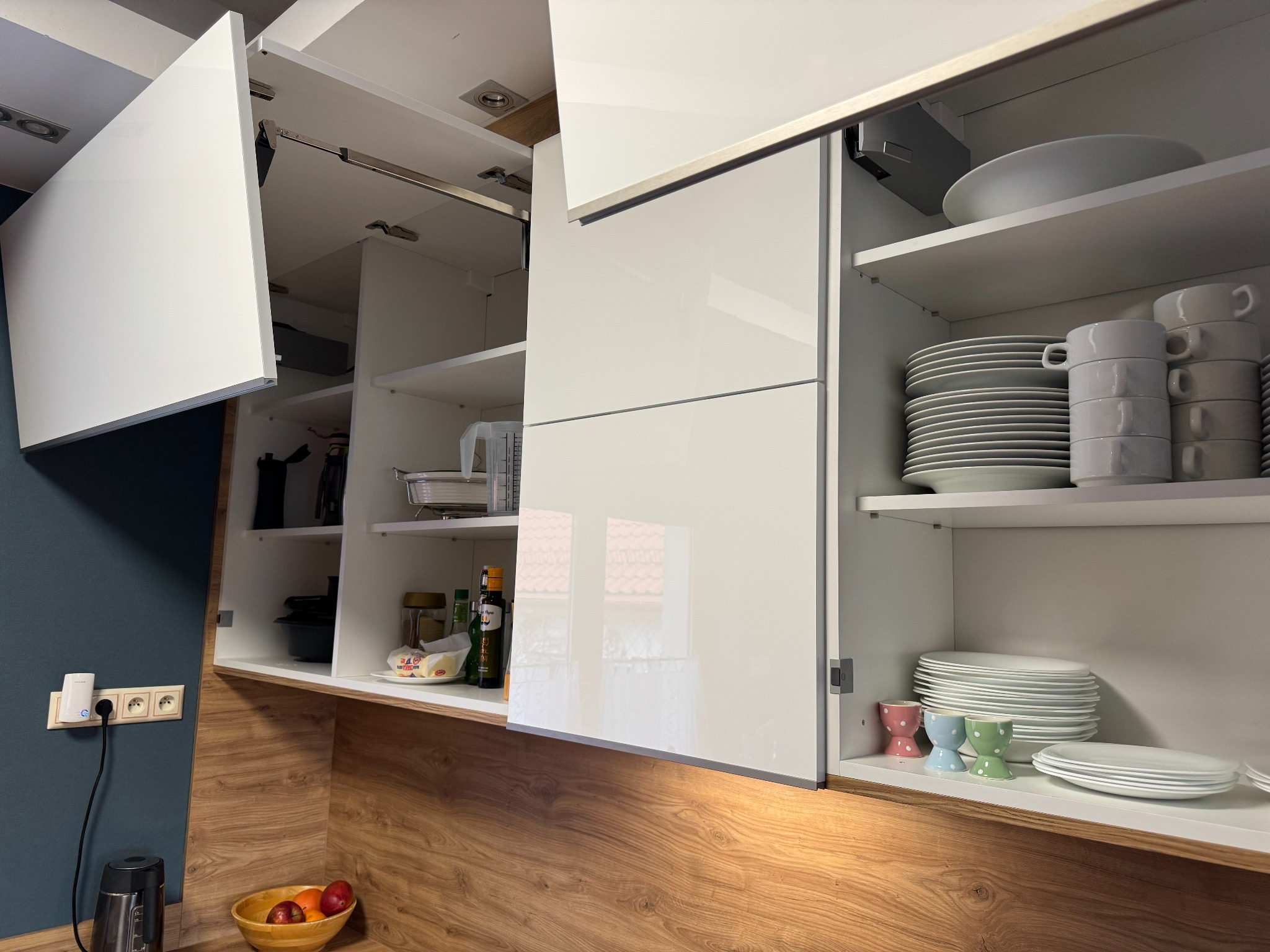 Nowoczesna kuchnia: białe, lakierowane fronty szafek otwarte, ukazujące zastawę. Blat w odcieniach drewna, na ścianie gniazdka elektryczne.