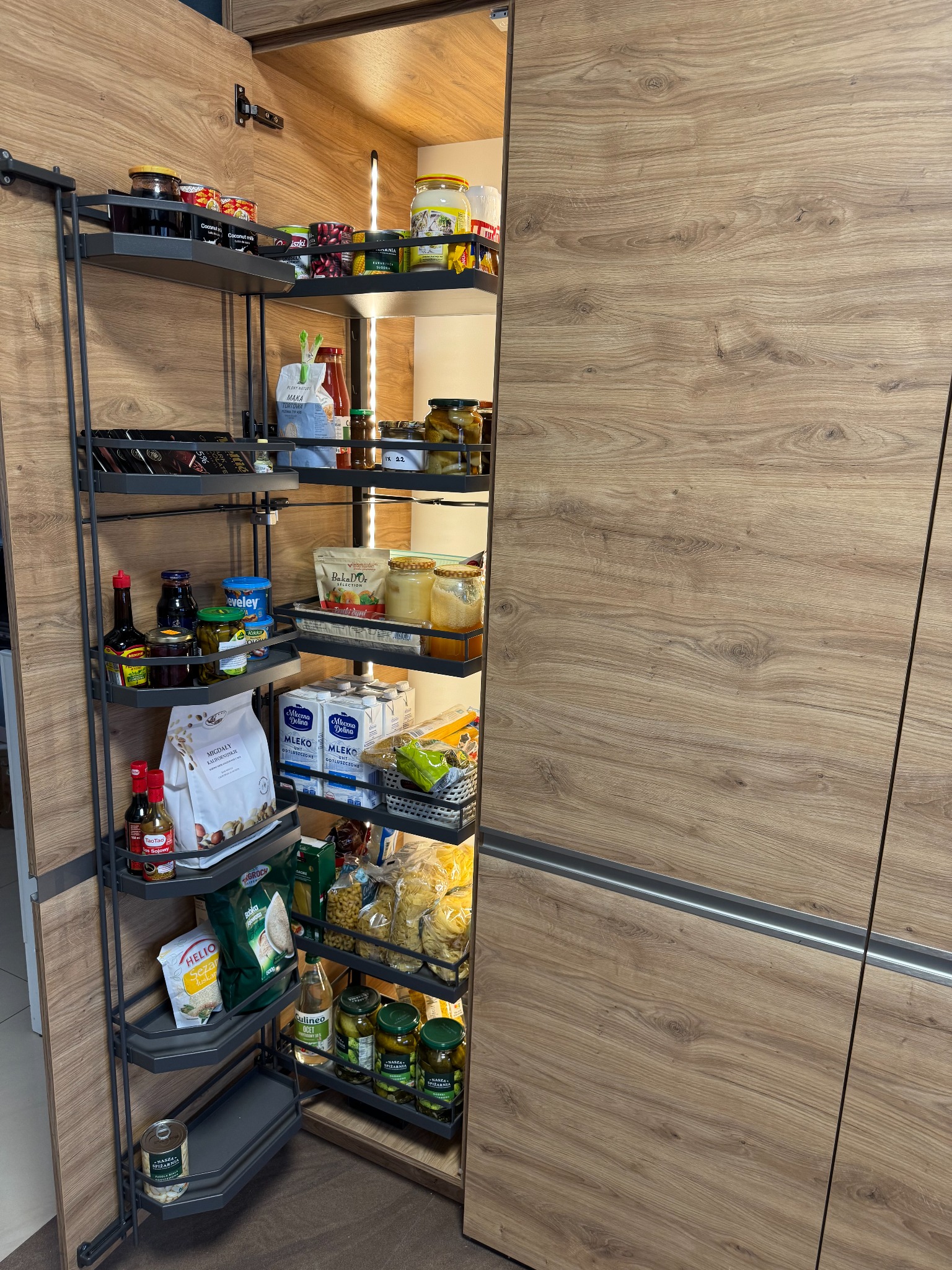 Drewniana szafa kuchenna z wysuwanymi półkami wypełnionymi produktami spożywczymi. Widoczne różne słoiki, puszki, makarony i inne artykuły. Oświetlenie LED wewnątrz szafy.