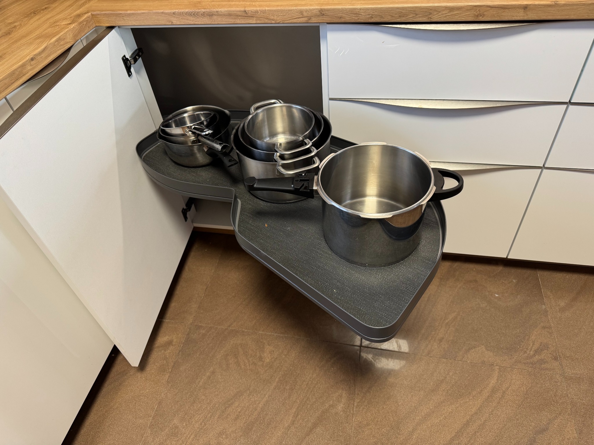 Funkcjonalna kuchnia na wymiar: wysuwana półka narożna z garnkami w szafce z białymi frontami i drewnianym blatem. Praktyczne rozwiązanie do przechowywania.