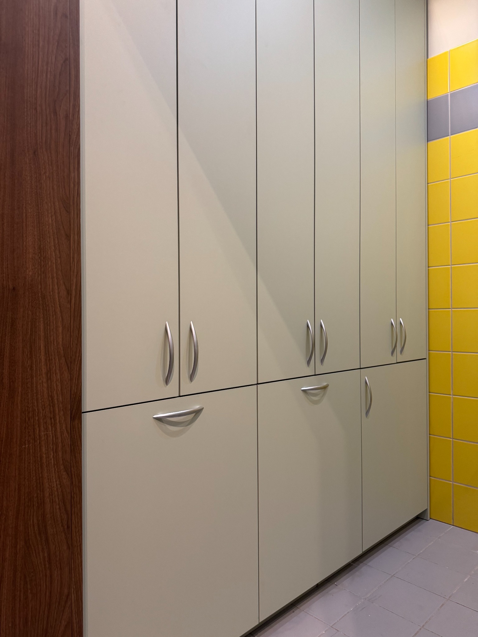 Białe szafy wnękowe z metalowymi uchwytami, drewniany bok i żółte płytki w tle. Proste, nowoczesne rozwiązanie do przechowywania, dopasowane do przestrzeni.