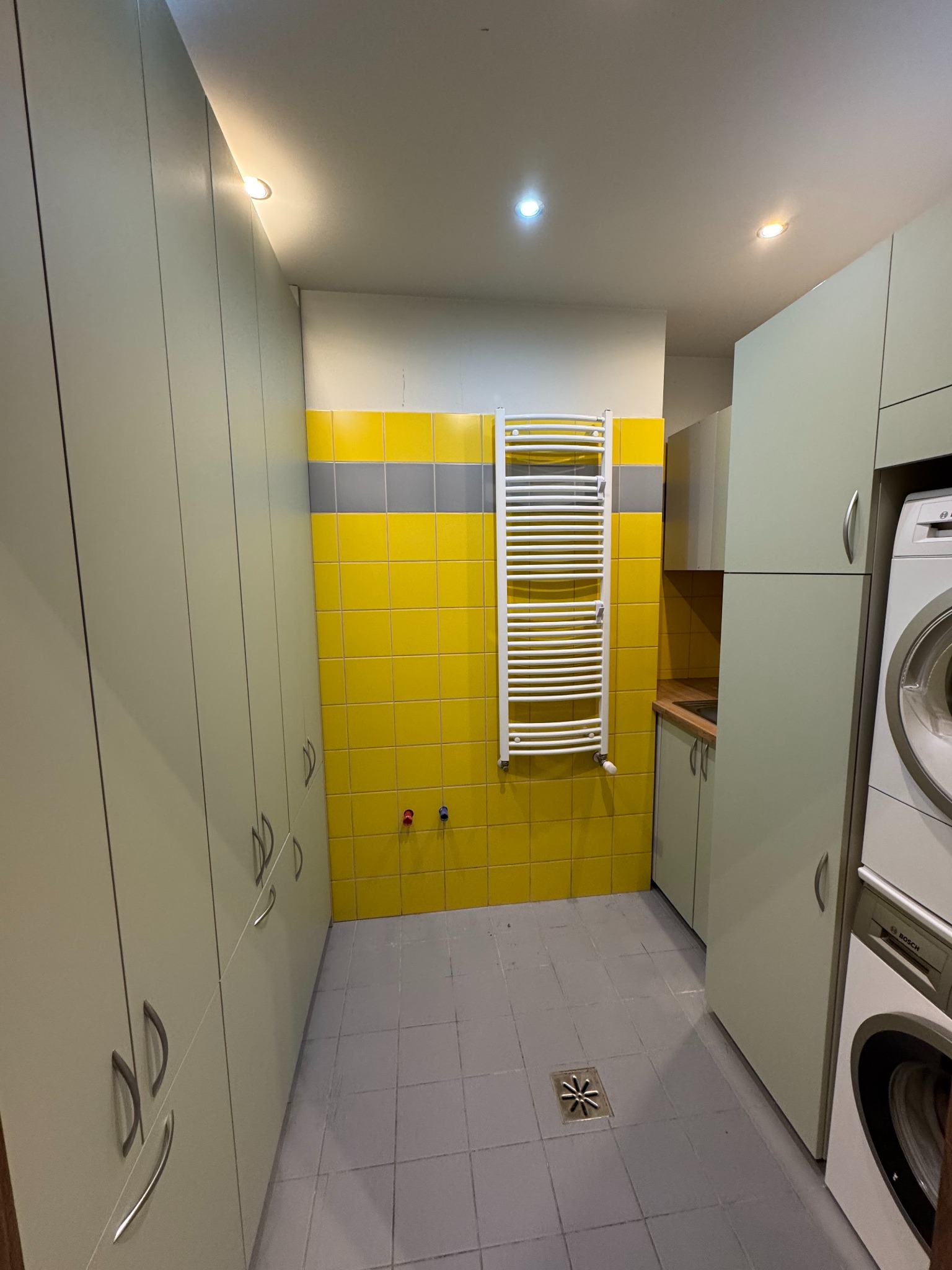 Funkcjonalna pralnia z szafami na wymiar w odcieniach szarości i żółci. Widoczna pralka Bosch, grzejnik drabinkowy i płytki ceramiczne na ścianie.
