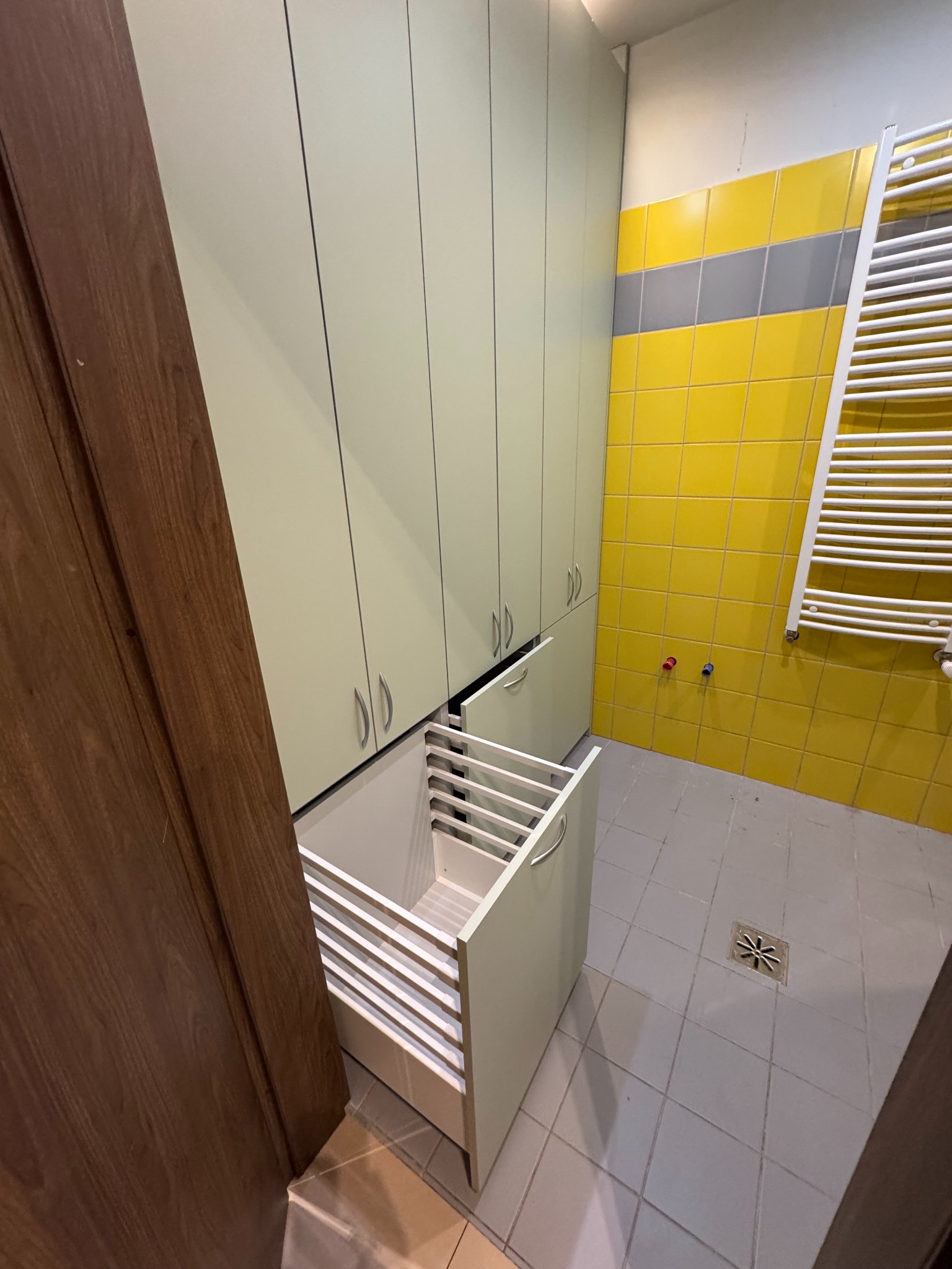 Wysoka szafa wnękowa z otwartą szufladą na pranie w łazience z żółtymi płytkami. Widoczne detale wykończenia i system szuflad.
