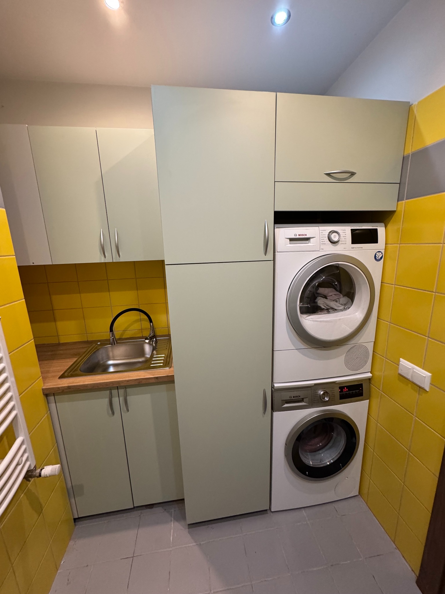 Jasnozielone szafki na wymiar z pralką i suszarką Bosch w żółtej łazience. Kompaktowe rozwiązanie do małych przestrzeni, nowoczesny design, praktyczne zagospodarowanie.