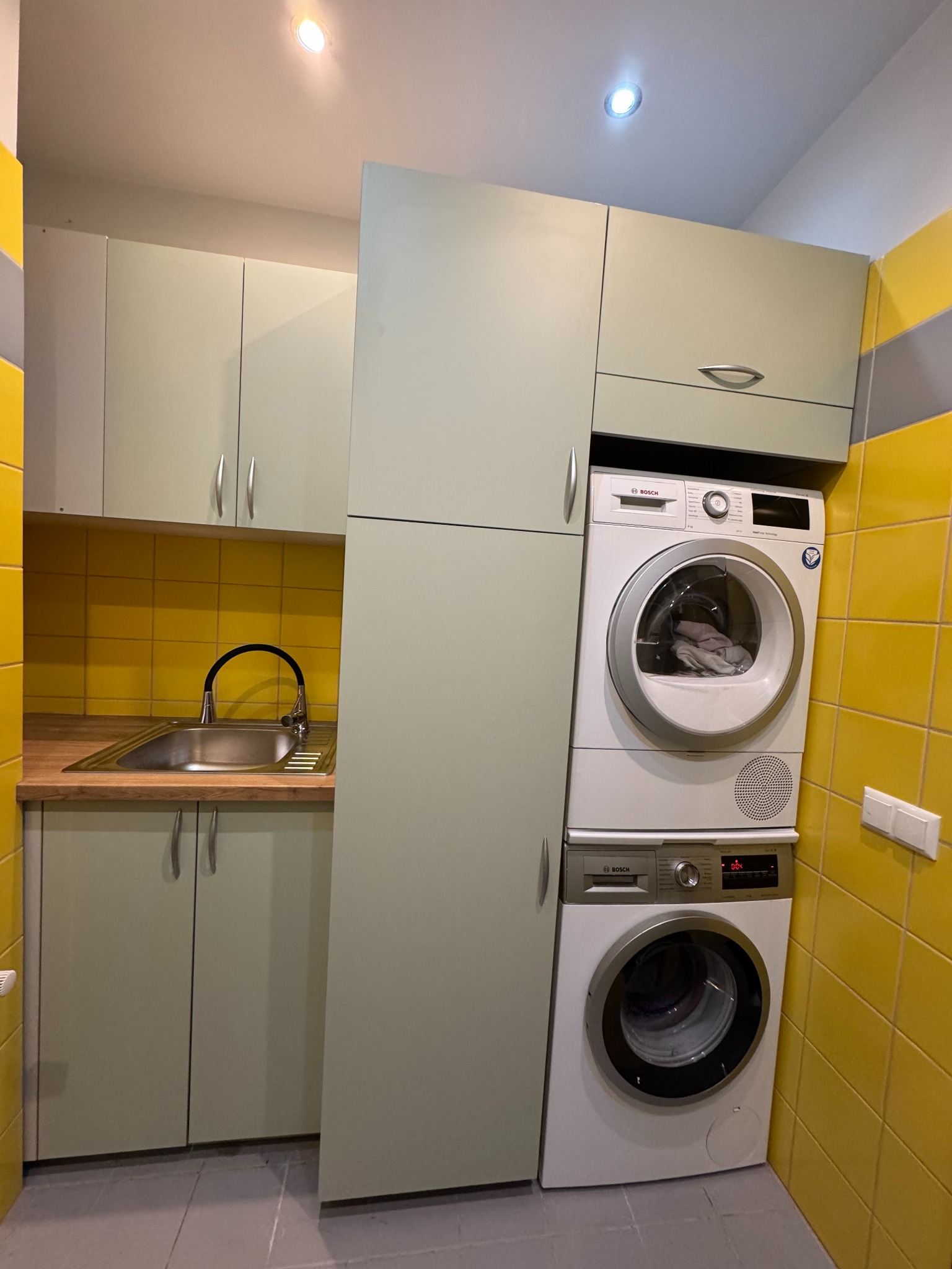 Narożna zabudowa kuchenna z pralką i suszarką Bosch w jasnej tonacji, żółte płytki na ścianie, szare płytki na podłodze, nowoczesny design.