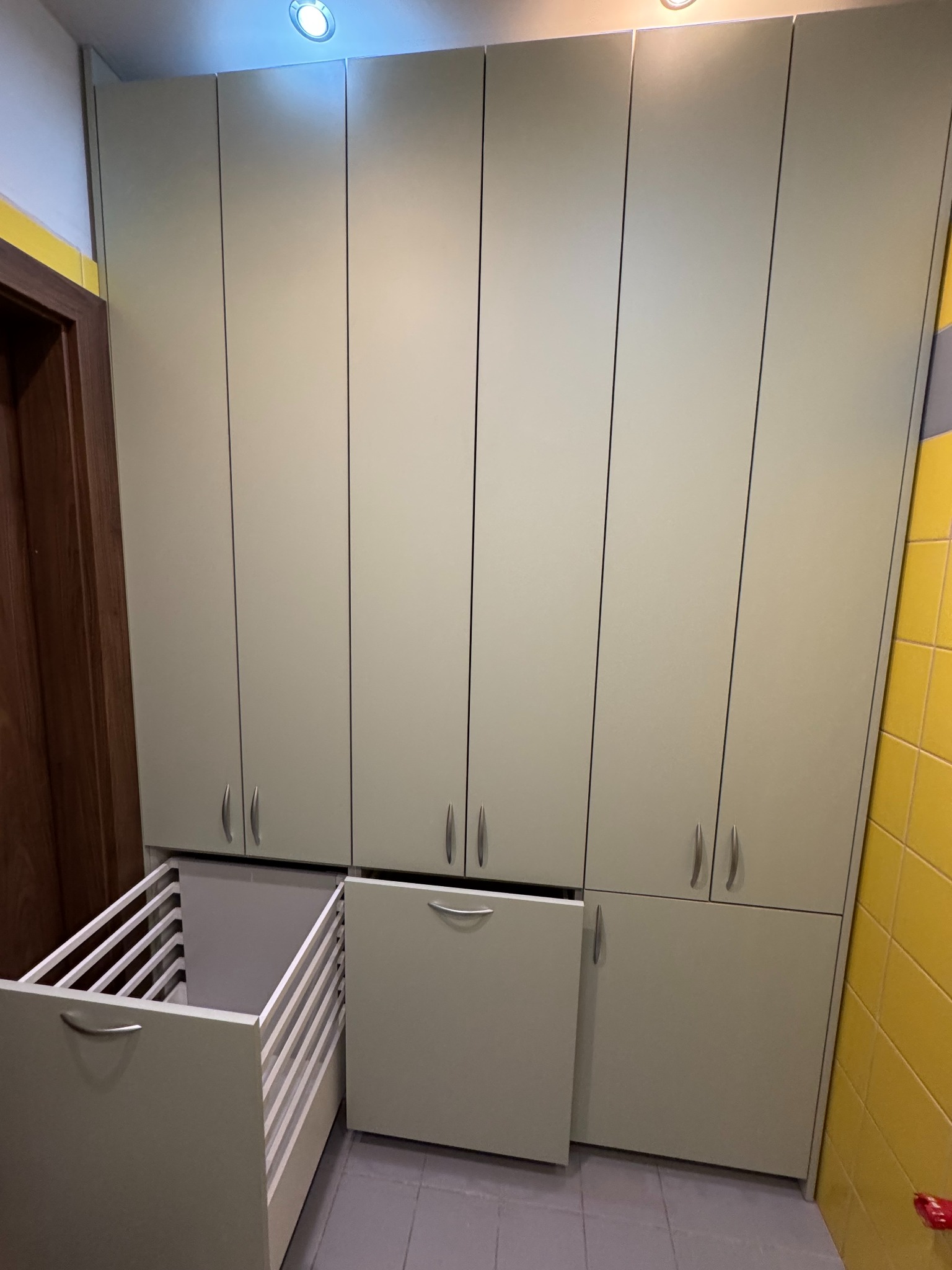 Szafa wnękowa z szufladą na pranie w Bagieńcu. Jasne fronty, minimalistyczne uchwyty. Szuflada z drewnianymi listwami, otwarta. Żółte płytki w tle.