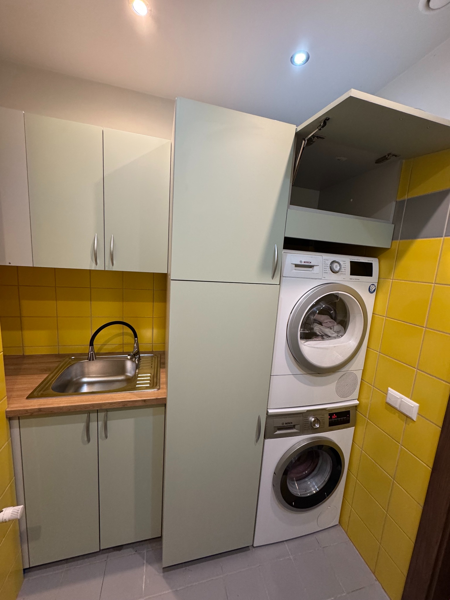 Funkcjonalna zabudowa: pralka i suszarka w szafie na wymiar z szafkami kuchennymi w pistacjowym odcieniu, żółte płytki na ścianie i szara podłoga.