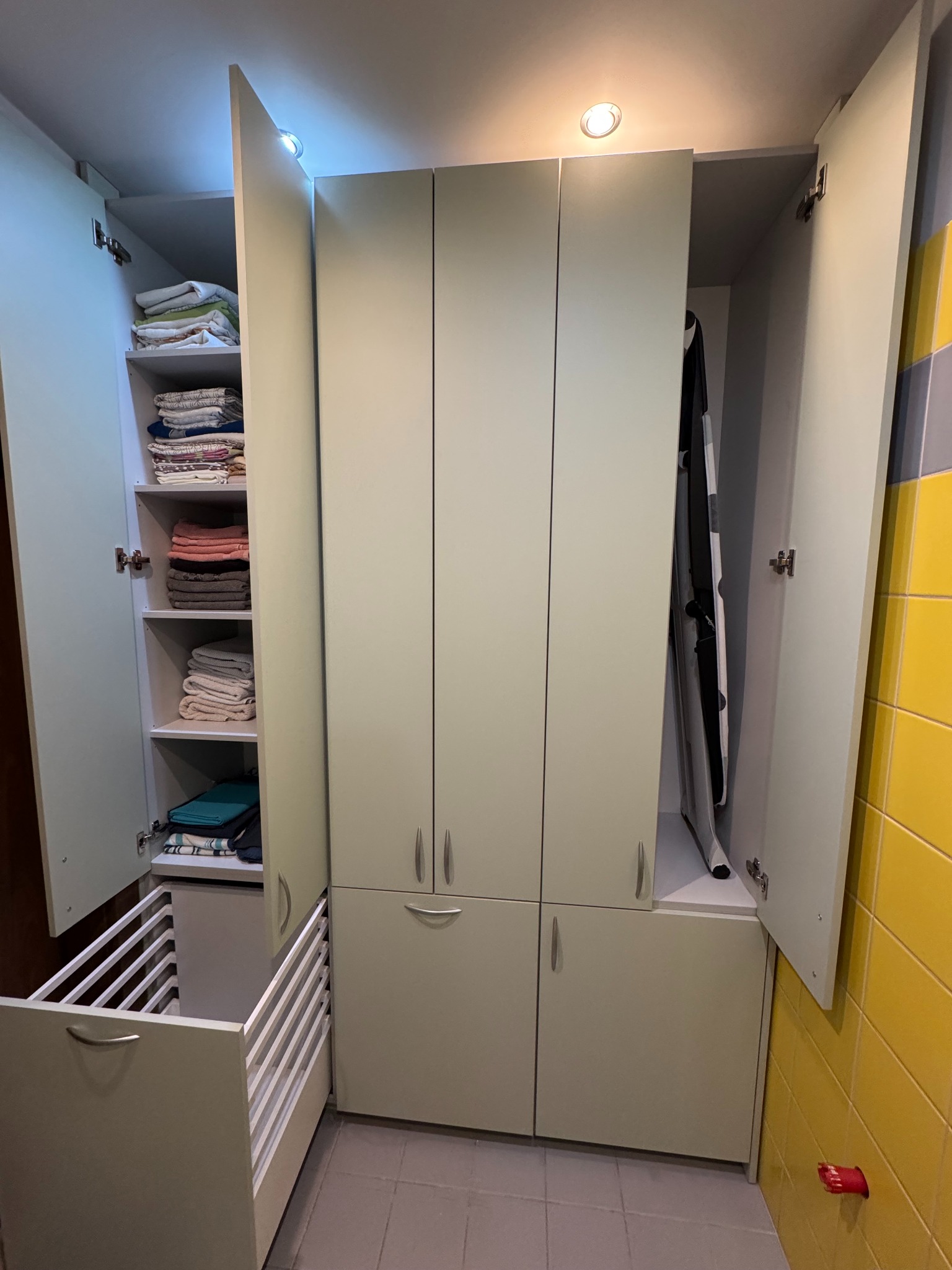 Jasna szafa na wymiar z otwartymi drzwiami, wewnątrz półki z ręcznikami i deską do prasowania. Dolna część z wysuwanym koszem. Żółto-szara ściana w tle.