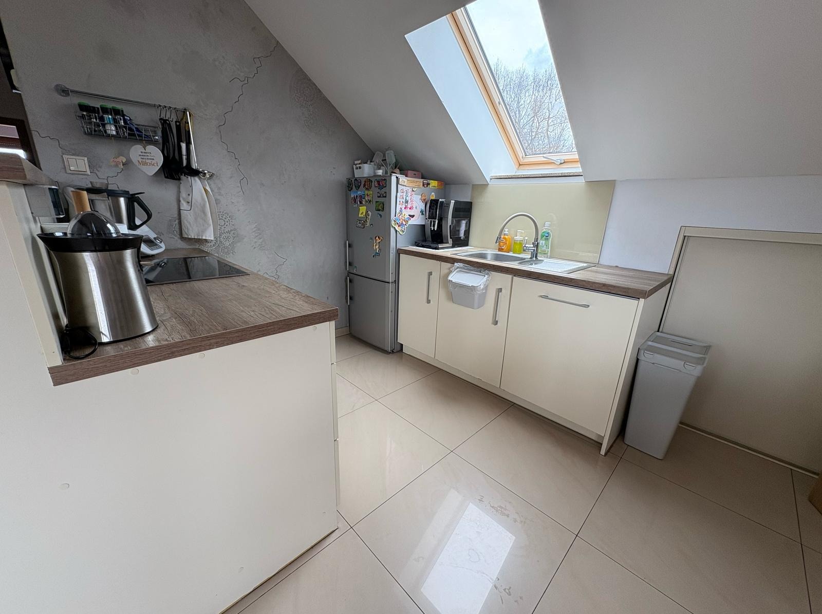 Jasna kuchnia na poddaszu z oknem dachowym. Kremowe szafki, drewniany blat, srebrna lodówka z magnesami i szara ściana z dekoracyjną fakturą.