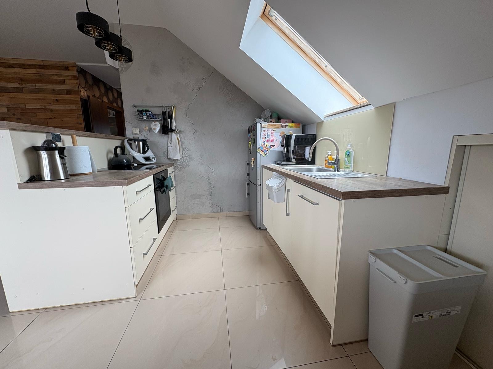 Jasna kuchnia na poddaszu z oknem dachowym. Zabudowa meblowa w odcieniach beżu z drewnianym blatem. Na blacie zlewozmywak i akcesoria kuchenne. Widoczny fragment lodówki i pojemnik.