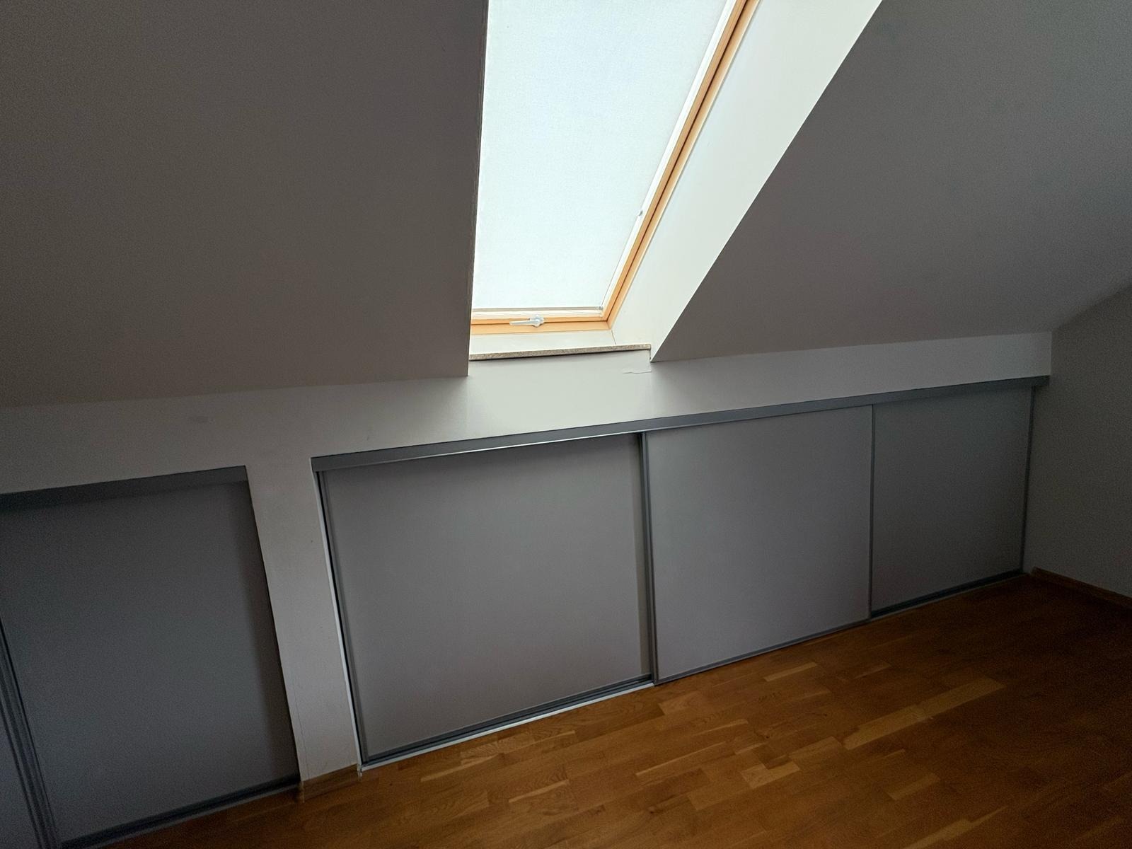 Szara szafa wnękowa z przesuwnymi drzwiami, wkomponowana w skos poddasza, oświetlona przez okno dachowe. Parkiet na podłodze. Minimalistyczny design.