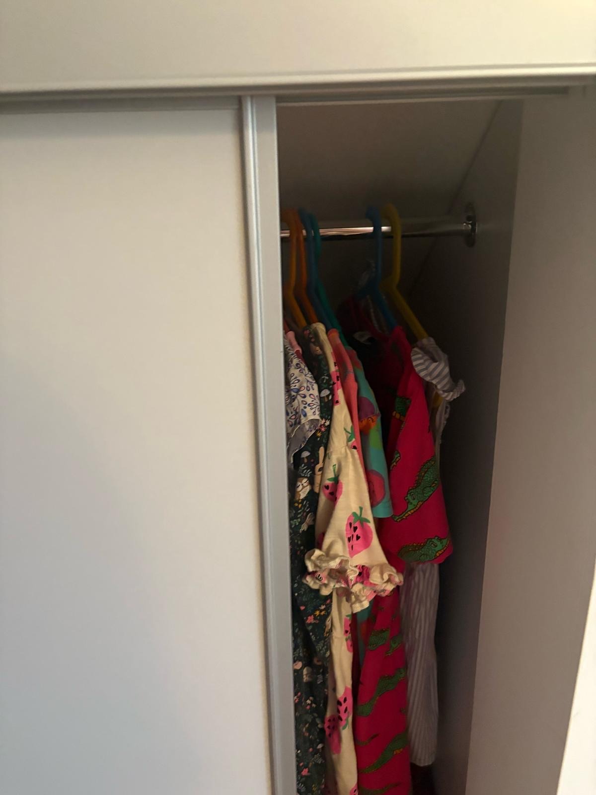 Biała szafa przesuwna z kolorowymi ubraniami na wieszakach. Drzwi szafy częściowo otwarte, ukazujące zawartość. Wnętrze oświetlone, widoczne detale ubrań.