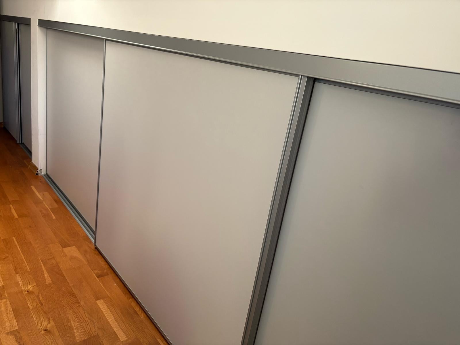 Nowoczesna szafa wnękowa z przesuwnymi, matowymi drzwiami w kolorze szarym. Widoczny fragment drewnianej podłogi i metalowe prowadnice. Minimalistyczny design.