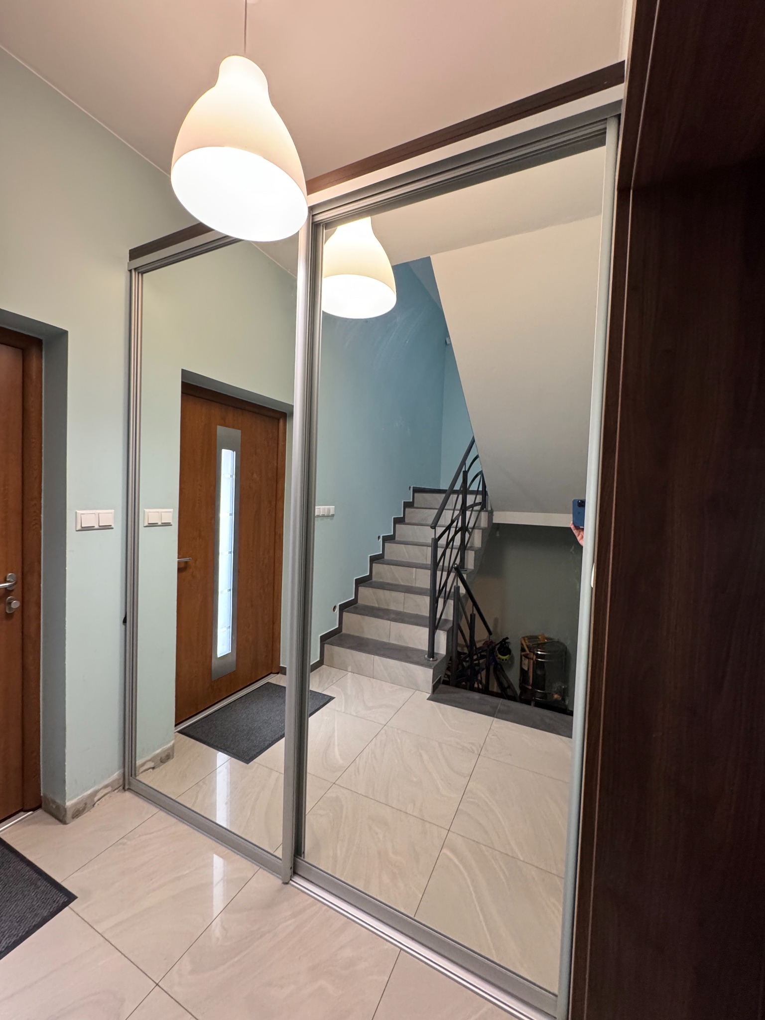 Szafa wnękowa z przesuwnymi lustrzanymi drzwiami w przedpokoju z widokiem na schody, oświetlona lampą sufitową. Widoczne wykończenie w kolorze ciemnego drewna.