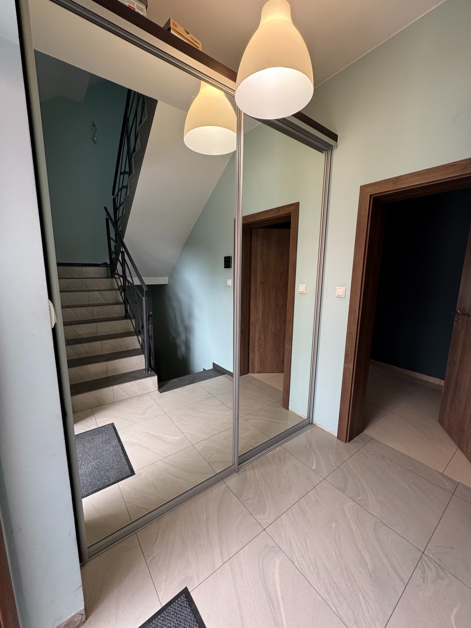 Szafa wnękowa z lustrzanymi drzwiami przesuwnymi w przedpokoju, odbijająca schody i lampę sufitową. Widoczne dwie pary drzwi po bokach szafy.