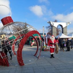 KROKODYLEK - Świąteczny jarmark miejski: Święty Mikołaj z workiem prezentów stoi obok dużej, dekoracyjnej bombki na tle straganów i pomnika pod błękitnym niebem.