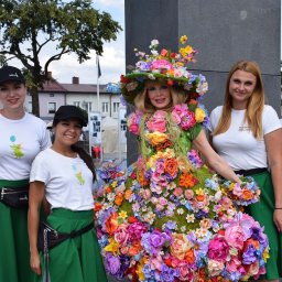 KROKODYLEK - Grupa kobiet w barwnych strojach kwiatowych, prezentujących się na tle miejskiego placu. Uwagę przykuwa centralna postać w sukni z żywych kwiatów.