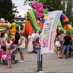 KROKODYLEK - Festyn rodzinny z dmuchańcami: zamek w kształcie zoo z małpą, zjeżdżalnia ze słoniem, tłum ludzi i baner reklamowy z ofertą animacji, waty cukrowej i fotobudki.