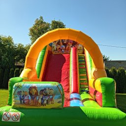 KROKODYLEK - Kolorowa dmuchana zjeżdżalnia z motywem zwierząt na trawie w słoneczny dzień. Widoczne drzewa w tle. Atrakcja dla dzieci na imprezę plenerową.