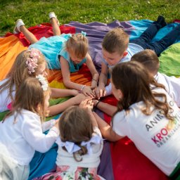 KROKODYLEK - Grupa dzieci w strojach leży na tęczowym kocu, trzymając się za ręce. Widoczna koszulka z napisem 'Animoksy'.