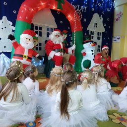 KROKODYLEK - Świąteczna impreza dla dzieci: Mikołaj w otoczeniu dmuchanych dekoracji i dzieci przebranych za aniołki, siedzących na dywanie. W tle dekoracje świąteczne.