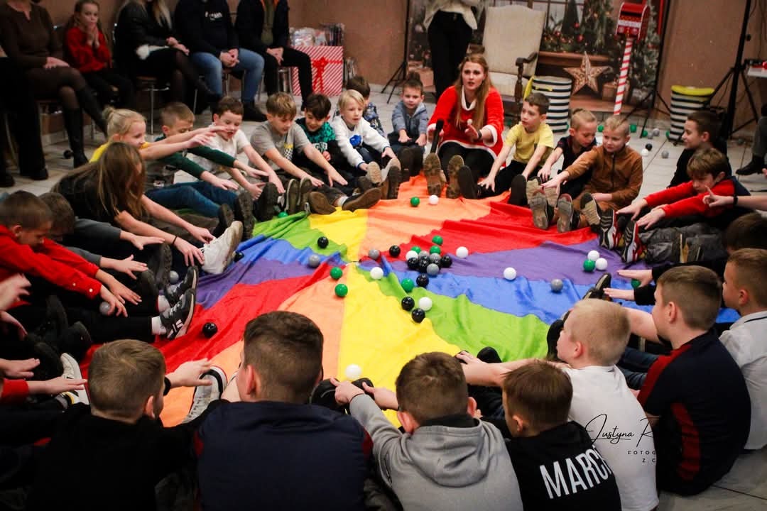 Grupka dzieci siedzi wokół tęczowej chusty animacyjnej z piłeczkami, prowadzona przez animatorkę w czerwonym stroju. Sala z dekoracjami świątecznymi w tle.