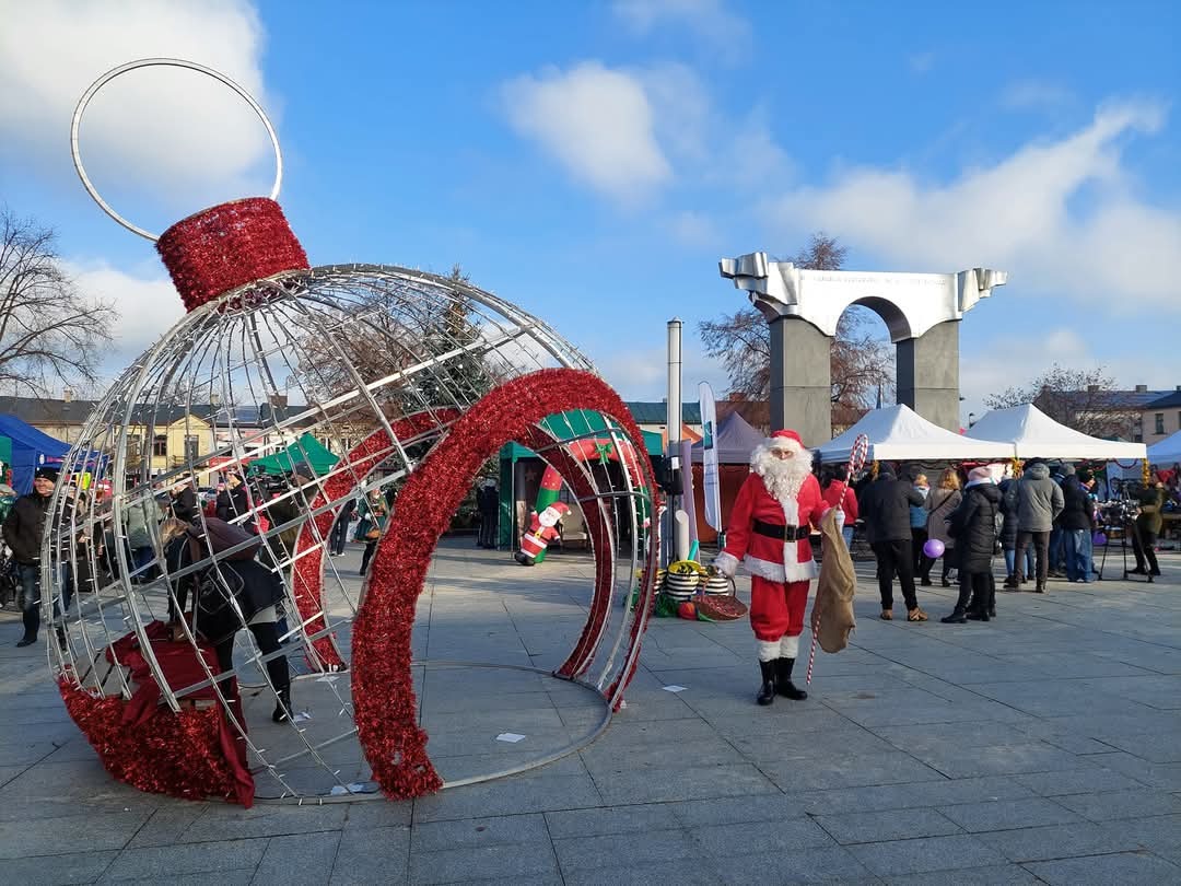 Świąteczny jarmark miejski: Święty Mikołaj z workiem prezentów stoi obok dużej, dekoracyjnej bombki na tle straganów i pomnika pod błękitnym niebem.