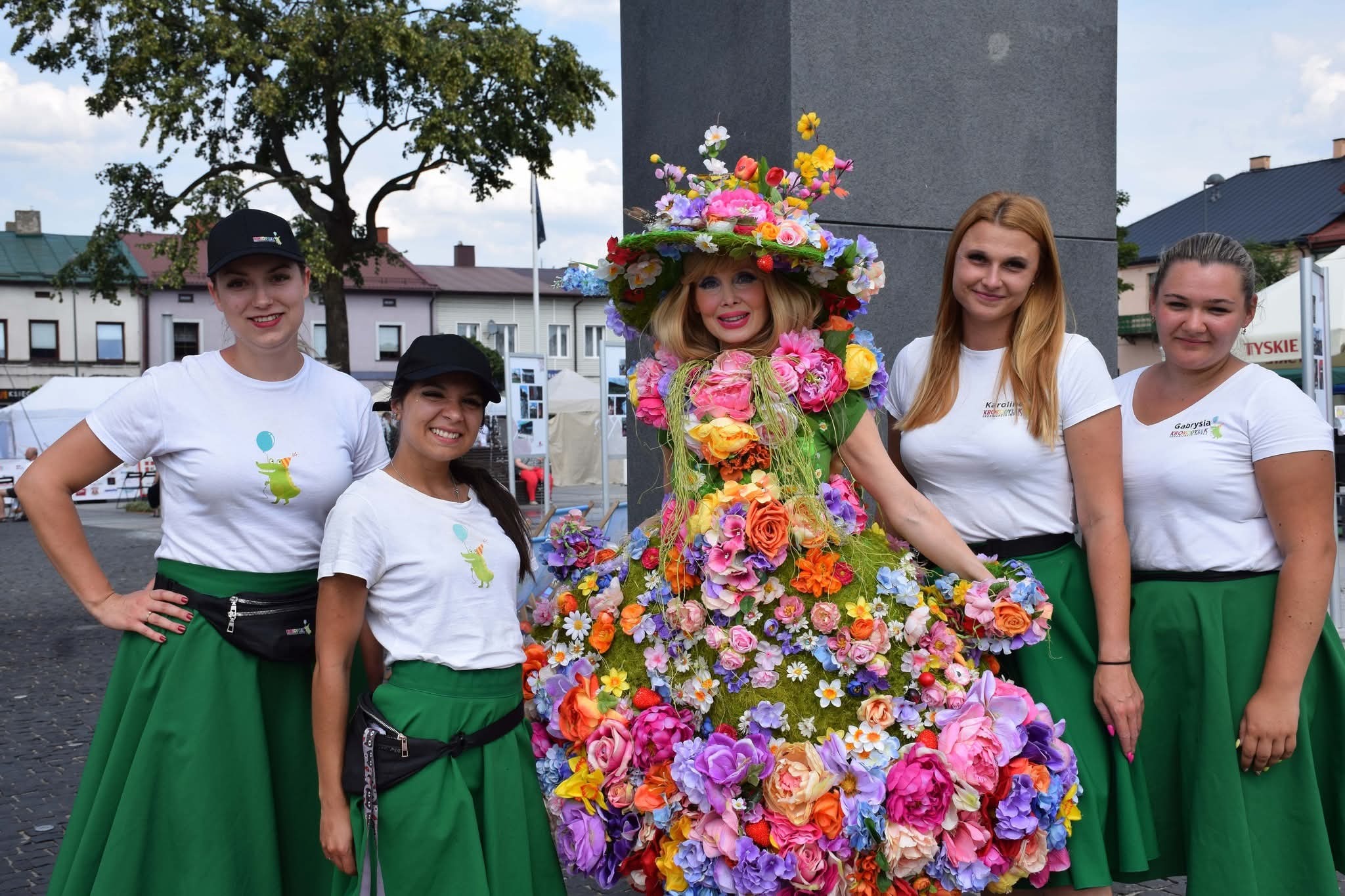 Grupa kobiet w barwnych strojach kwiatowych, prezentujących się na tle miejskiego placu. Uwagę przykuwa centralna postać w sukni z żywych kwiatów.