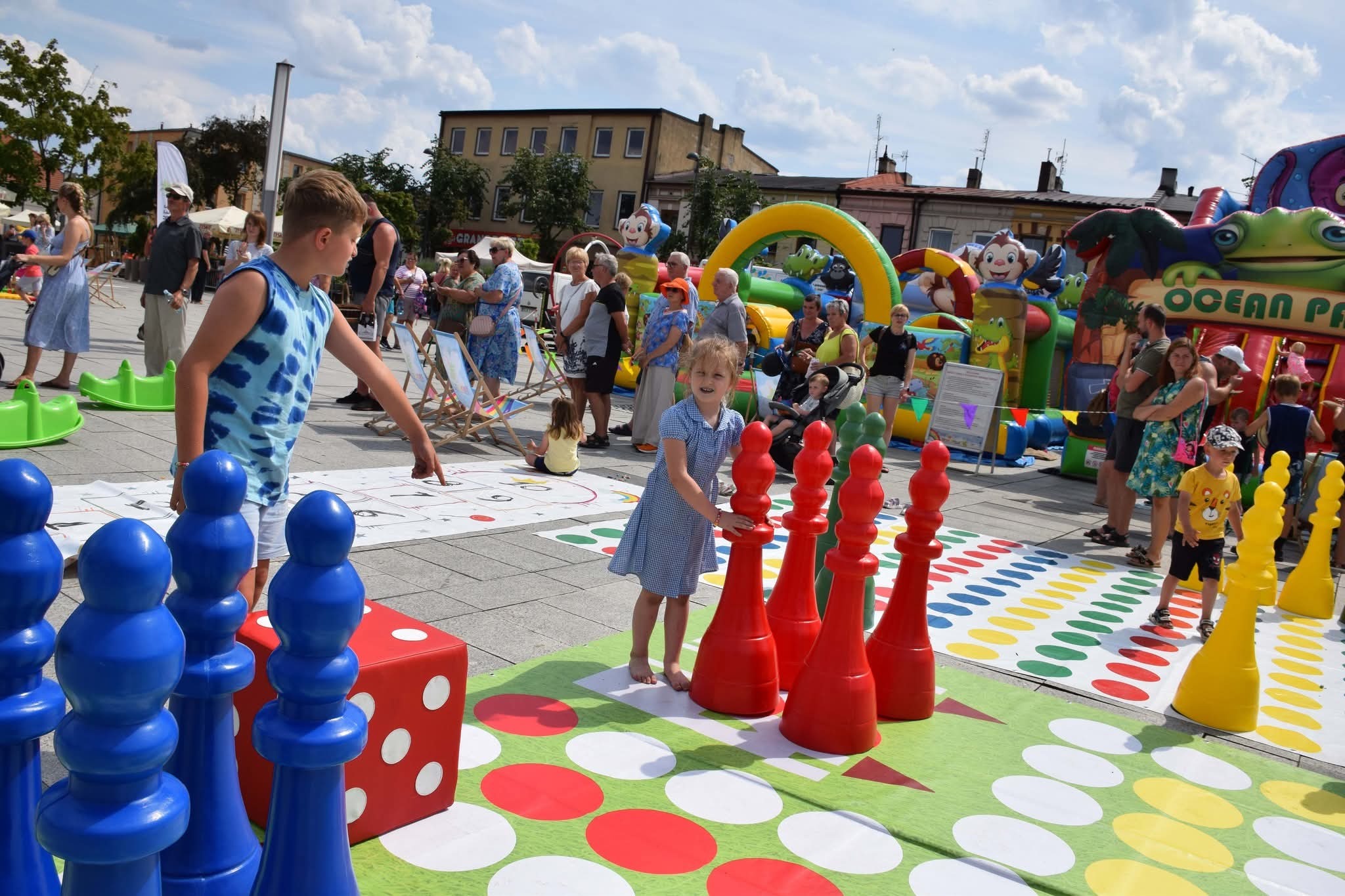 Plenerowa gra planszowa z dużymi pionkami i kostką, dzieci grają na placu miejskim, w tle dmuchany zamek i zgromadzeni ludzie.