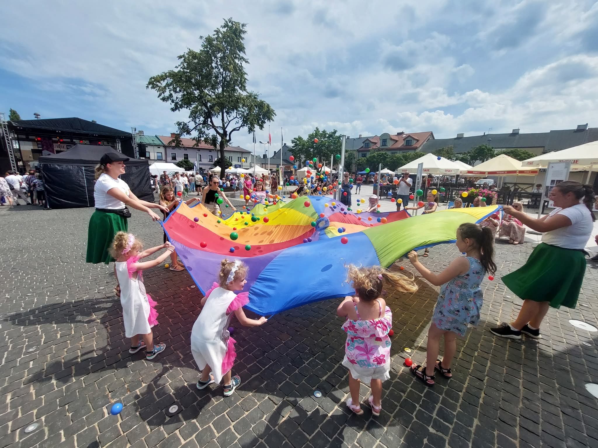 Kolorowa chusta animacyjna z piłeczkami w rękach dzieci i animatorów na tle miejskiego placu podczas imprezy plenerowej.