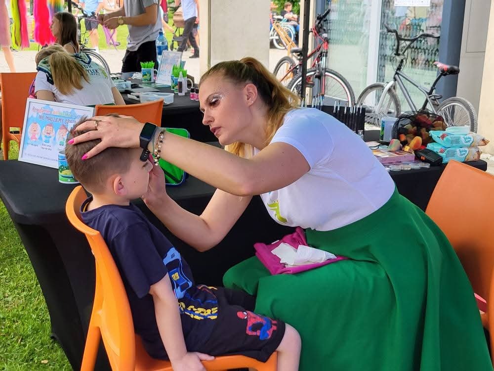 Animator maluje twarz chłopcu na krześle. Widoczne farby, pędzle i akcesoria. W tle rowery i przechodnie. Kobieta ma brokatowy makijaż.