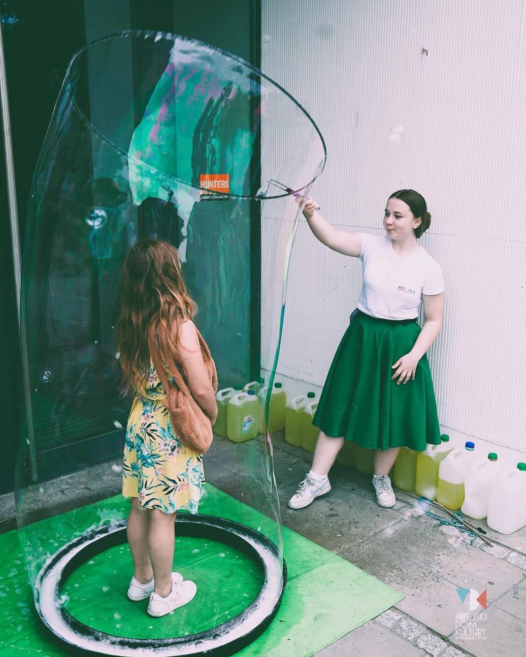 Dziewczynka wewnątrz gigantycznej bańki mydlanej, tworzonej przez animatorkę w zielonej spódnicy. Atrakcja plenerowa z płynem do baniek w tle.