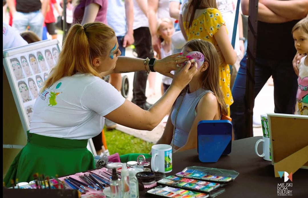 Malowanie twarzy dziewczynce przez animatorkę z brokatowym makijażem, na tle tłumu i tablicy z wzorami, na stole pędzle i farby. Usługa na imprezie plenerowej.