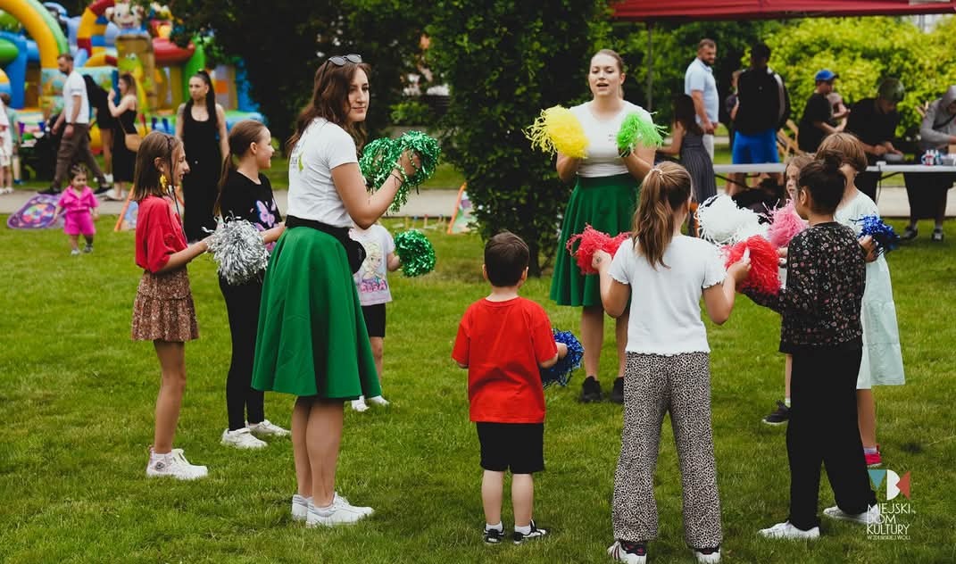 Animatorki w zielonych spódnicach z kolorowymi pomponami prowadzą animacje dla dzieci na trawie podczas imprezy plenerowej. W tle dmuchany zamek i inne atrakcje.