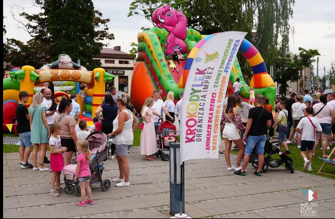Festyn rodzinny z dmuchańcami: zamek w kształcie zoo z małpą, zjeżdżalnia ze słoniem, tłum ludzi i baner reklamowy z ofertą animacji, waty cukrowej i fotobudki.