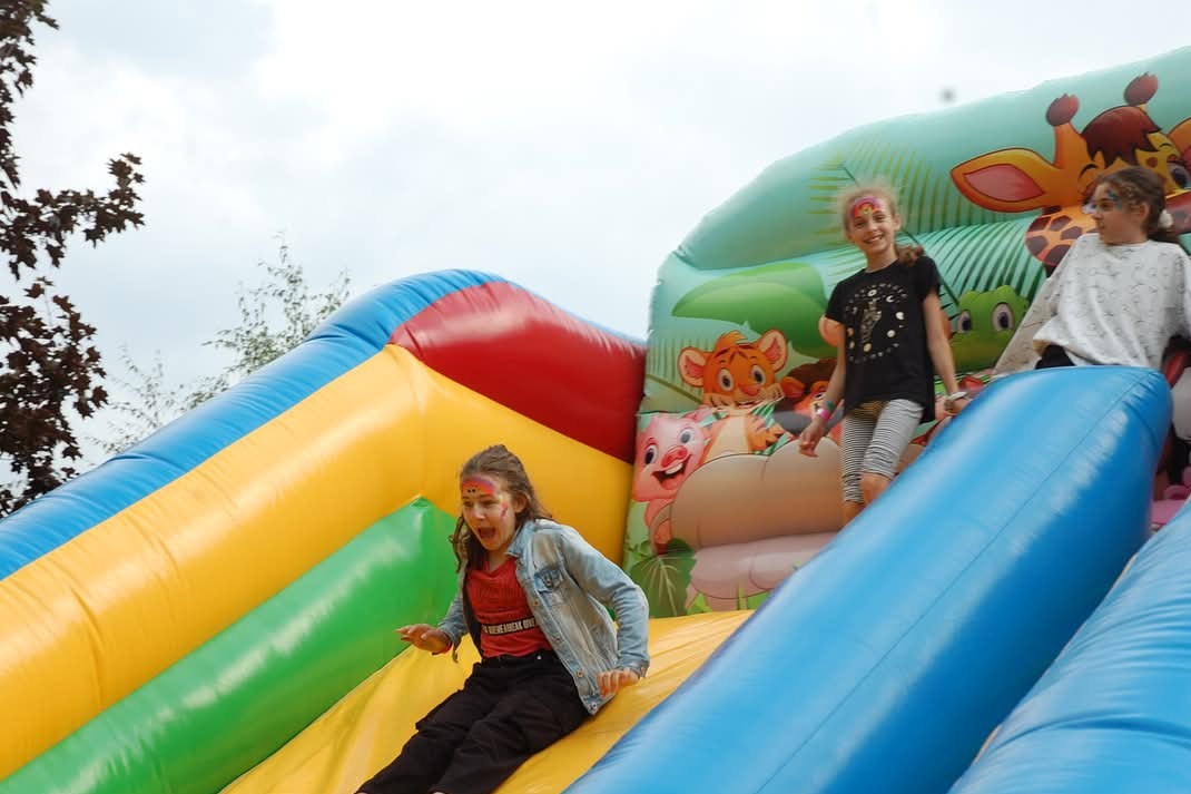 Kolorowa dmuchana zjeżdżalnia z motywem zwierząt, na której bawią się dzieci z pomalowanymi twarzami. Widok z boku, w tle drzewa i pochmurne niebo.