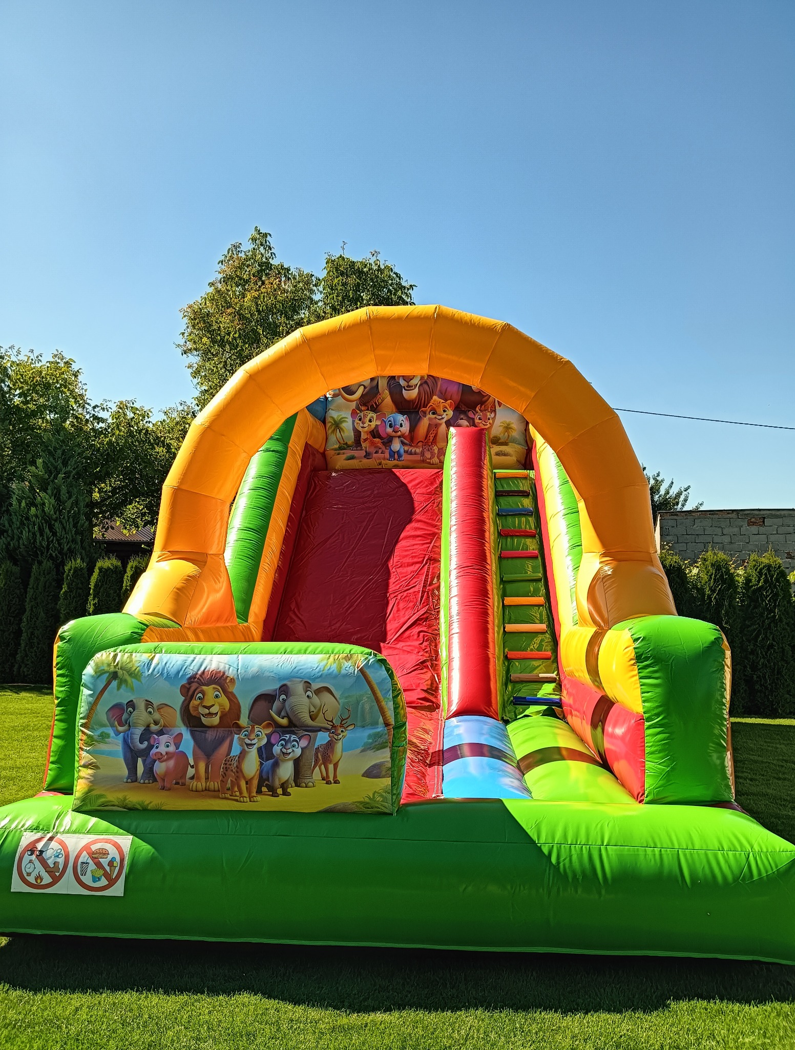 Kolorowa dmuchana zjeżdżalnia z motywem zwierząt na trawie w słoneczny dzień. Widoczne drzewa w tle. Atrakcja dla dzieci na imprezę plenerową.